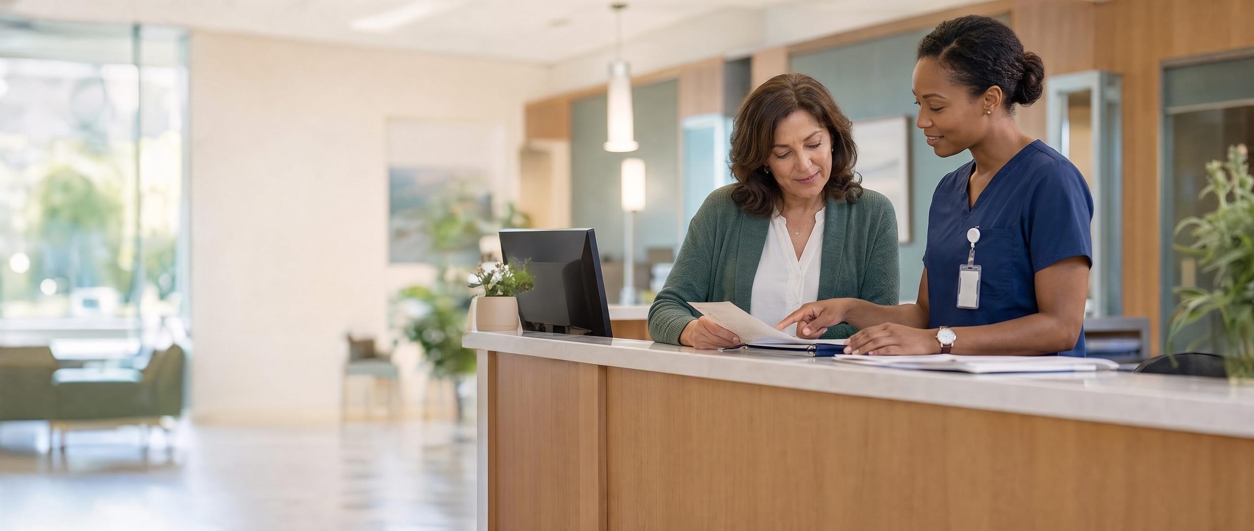 A front-desk coordinator and an African American nurse in navy scrubs review a document together at the reception desk of a bright outpatient clinic.