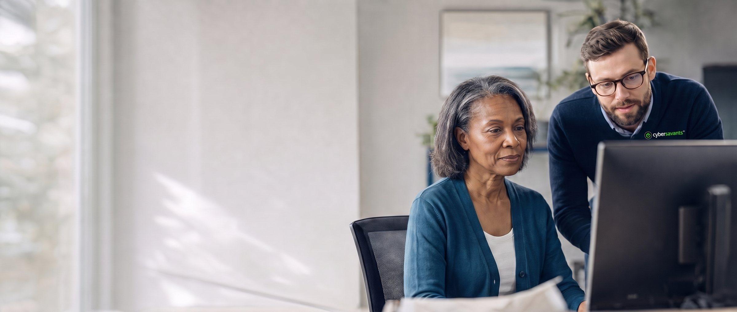 A practice administrator, an African American woman in her fifties, reviews a computer monitor with an IT specialist wearing a Cybersavants sweater, in a bright office.