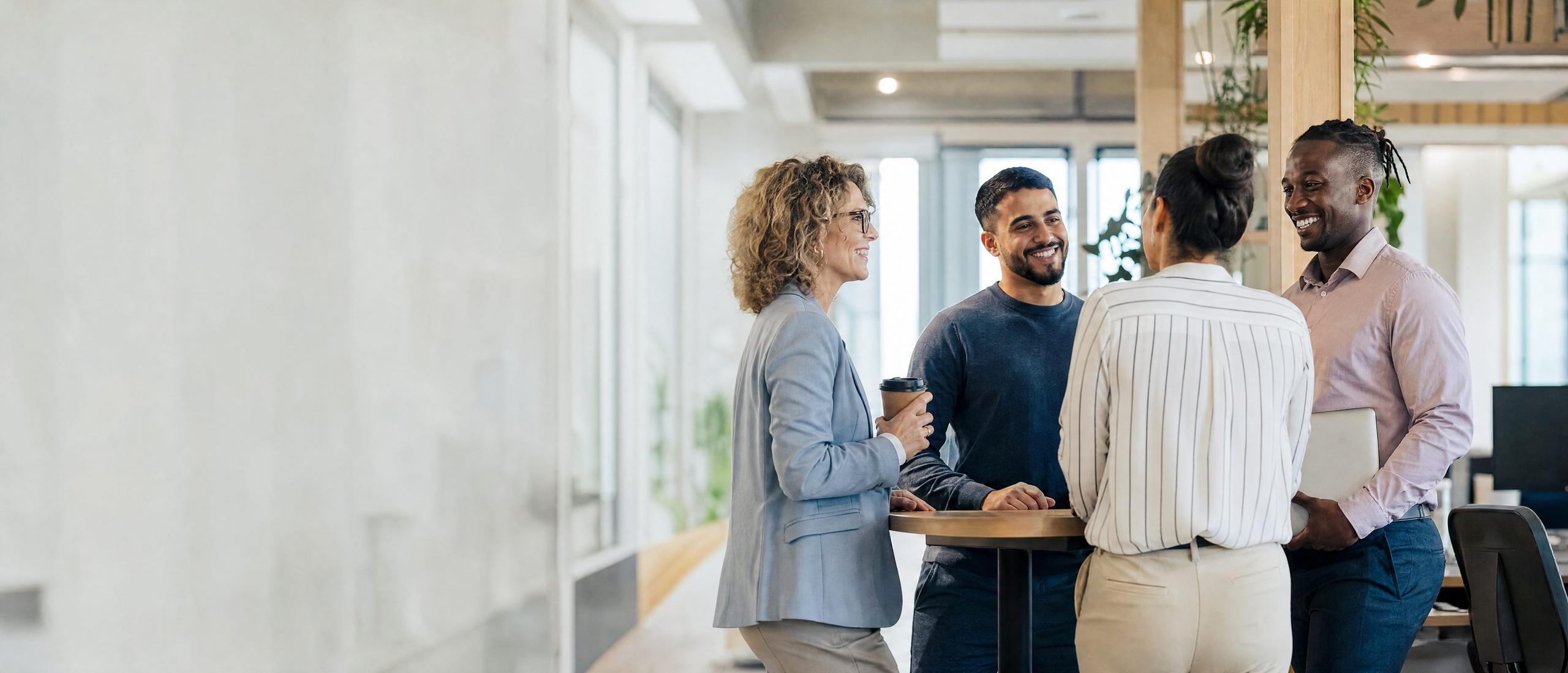 Four colleagues talk and laugh together around a standing table in a bright, modern office.